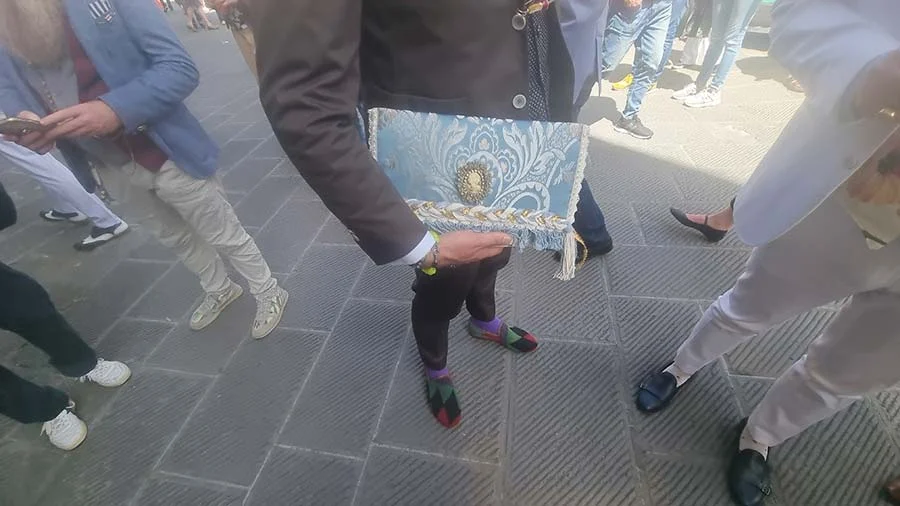 Pale blue brocade clutch with gold braid trim and a round clasp, carried by hand through the crowd