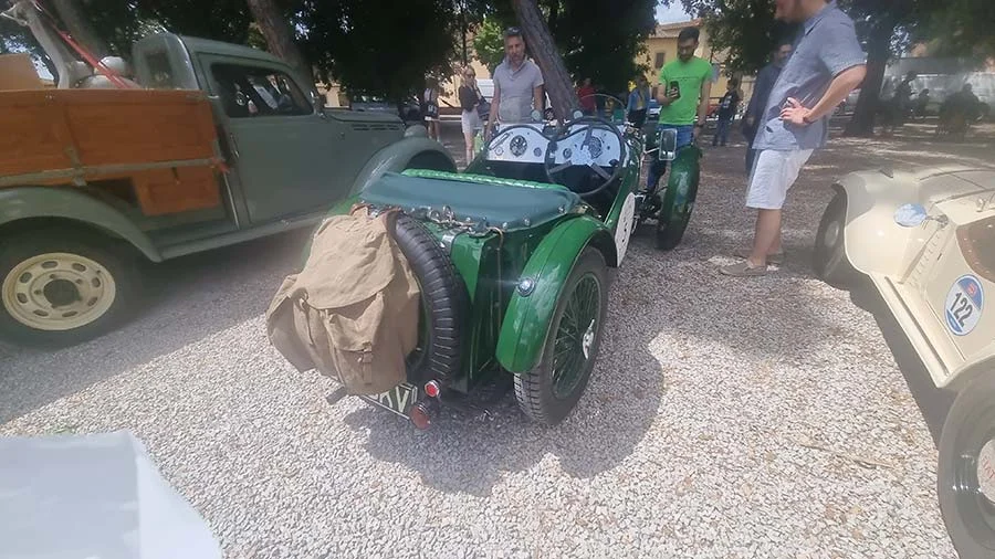 Vintage green open-top sports car with a spare wheel and a canvas bag strapped on the back, parked on gravel