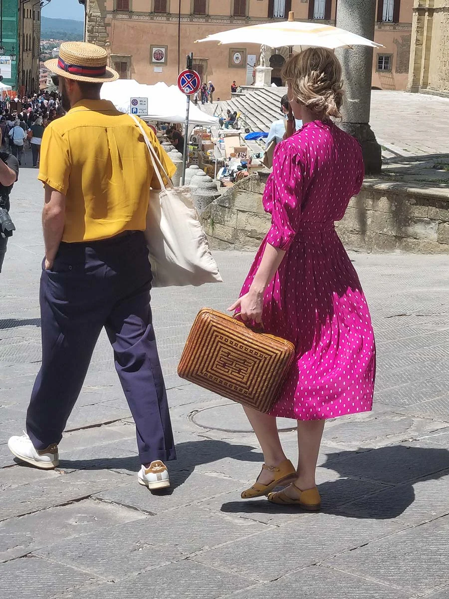 A woman in a magenta polka-dot dress carrying a woven basket bag, walking beside a man in a yellow shirt and straw hat