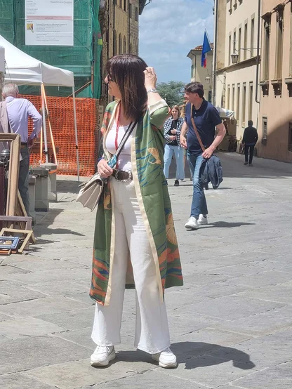 A woman in a long green and orange printed coat, white trousers and white trainers, with a cream shoulder bag