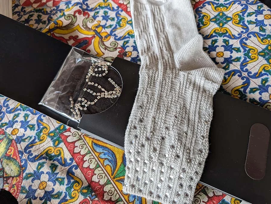 A grey knitted sock with studded grip beside a packet of black no-show socks with a crystal chain trim