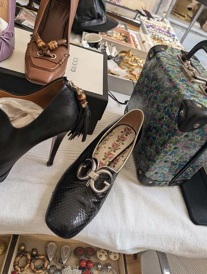 Black textured leather flat with a silver-tone clasp and floral insole, on a stall beside a Gucci shoe box