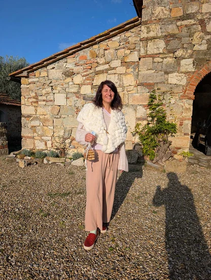 Woman in a fluffy white cropped jacket and pink trousers outside a stone farmhouse, holding a small woven bag