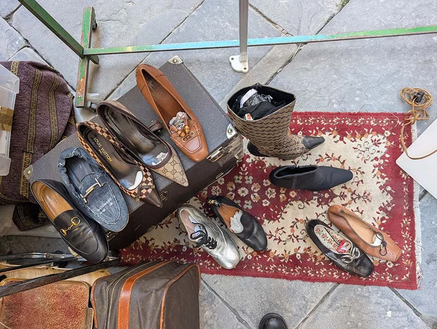 Second-hand loafers, pumps and a boot laid out on a patterned rug and an old suitcase at a market stall