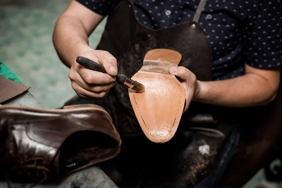 Cobbler in an apron brushing glue onto the tan leather sole of a shoe, a brown shoe beside him