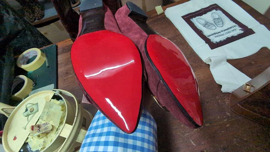 Pair of burgundy suede high heels turned over to show glossy red soles, on a workbench