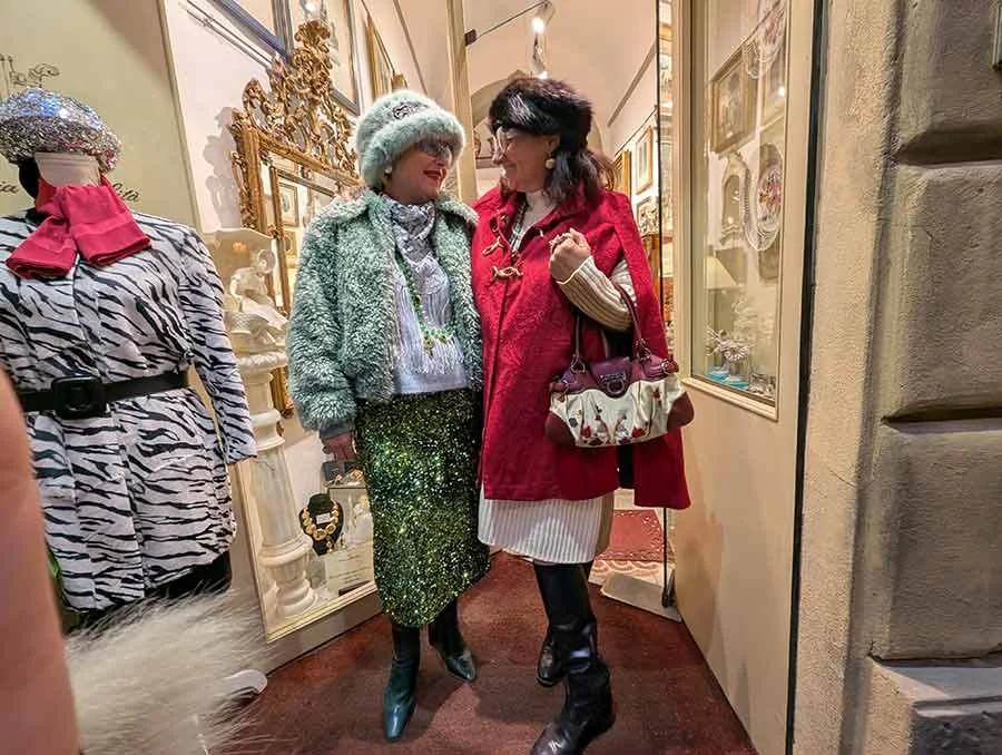 Two women in a boutique doorway: one in a green fur hat and jacket and sequinned skirt, one in a red coat with a floral bag