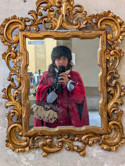 Gracie Opulanza photographed in a gilded mirror in a black fur hat and red coat, with a patterned handbag over her arm