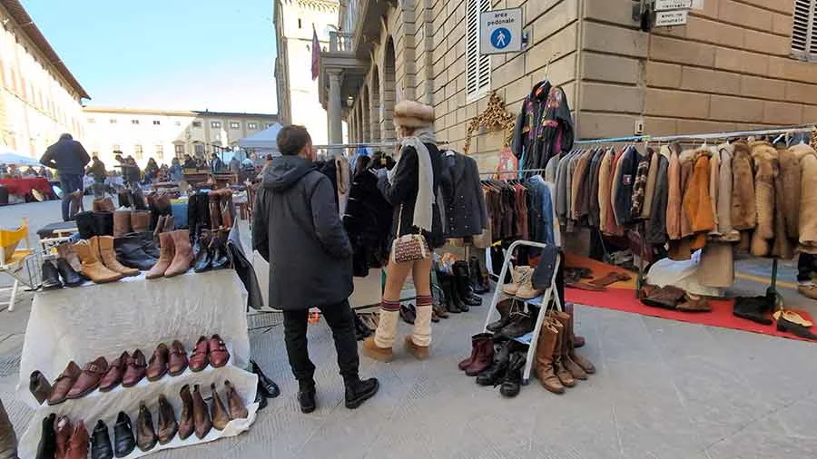 Street market with tables of leather boots and rails of coats, a woman in a fur hat and scarf among them