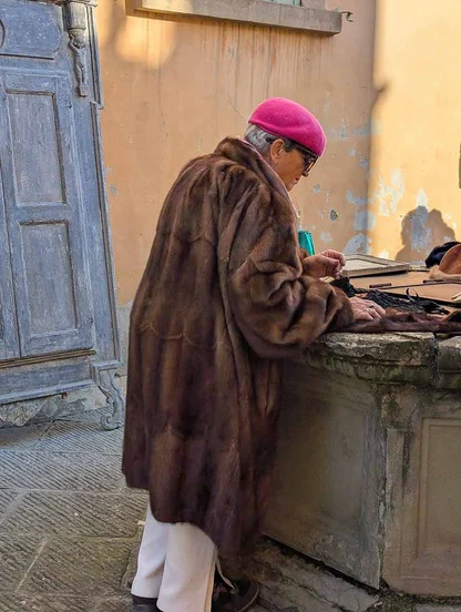 A woman in a pink beret and long brown mink coat looking over goods on a stone ledge, beside a weathered wooden door