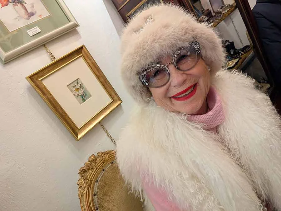 A smiling woman in a white fur hat and gilet with jewelled glasses, in a shop hung with framed pictures