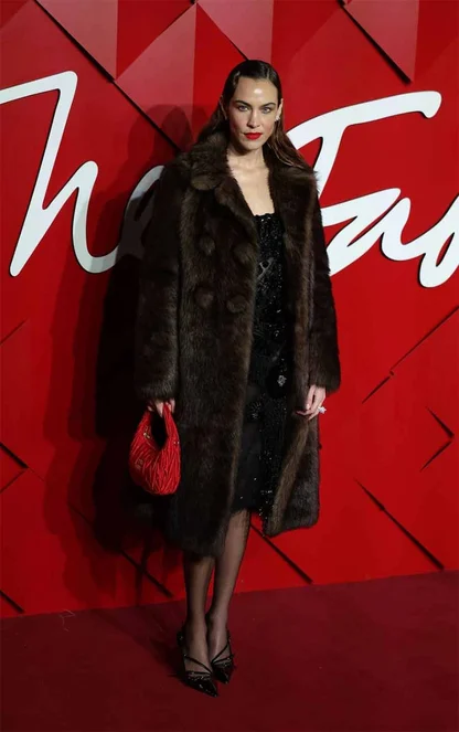 Alexa Chung in a long dark fur coat and black sequinned dress, with a red bag, against a red backdrop