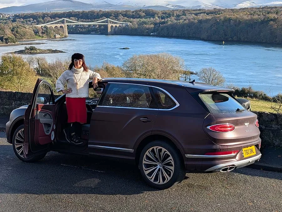 Gracie Opulanza in a mink hat, white knit and red skirt, leaning on a Bentley Bentayga with a bridge behind