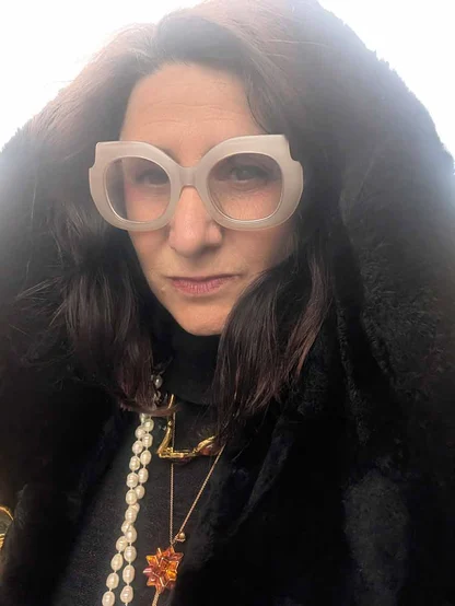 Close-up of Gracie Opulanza in oversized pale pink glasses with the faux fur hood up, wearing pearls and gold necklaces