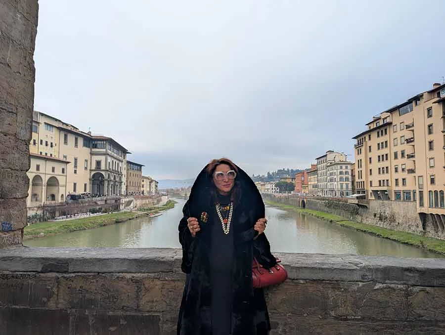 Black hooded faux fur cape worn with layered pearls and a red bag, with the Arno and Florence behind