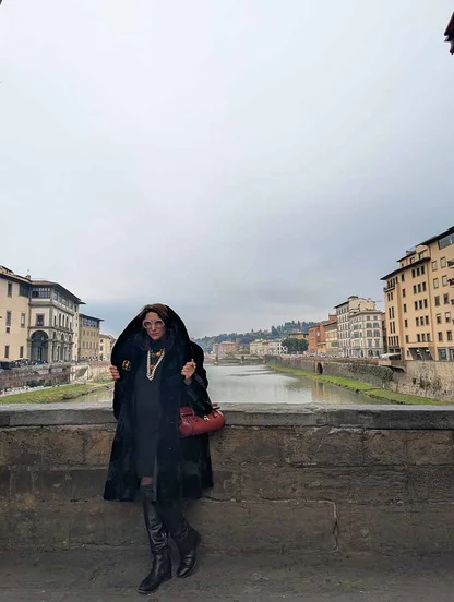 Full-length view on the Ponte Vecchio: black faux fur cape, long pearl necklace, red bag and black boots