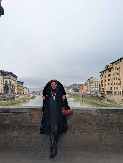 Gracie Opulanza on the Ponte Vecchio in Florence in a hooded black faux fur cape, with a red bag and knee-high black boots