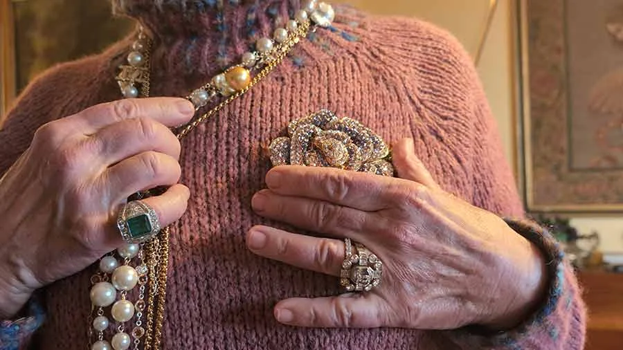 Layered pearl and gold necklaces, a large flower brooch and a green-stone ring worn over a pink knit jumper