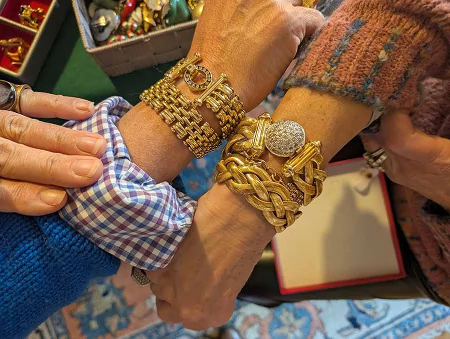Two wrists layered with chunky gold-tone bracelets, one braided with a round crystal-set clasp
