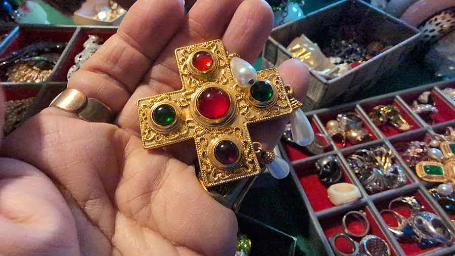 Gold-tone cross brooch set with red, green and dark stones and pearl drops, held in a hand above a tray of jewellery