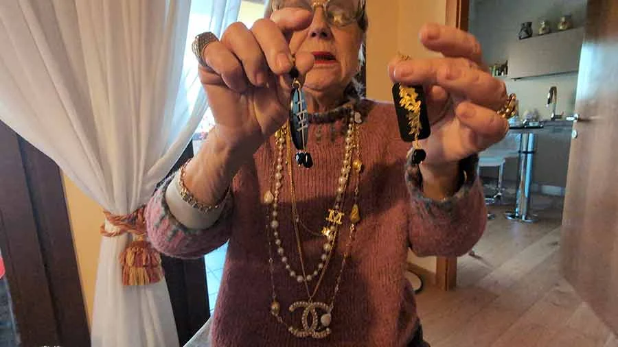 Woman holding up a pair of black and gold earrings, wearing layered pearl and gold chains with a Chanel pendant