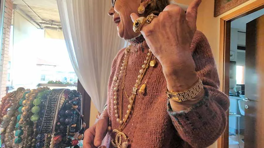 Older woman in a pink knit jumper wearing long gold chains and pearls with a Chanel double-C pendant