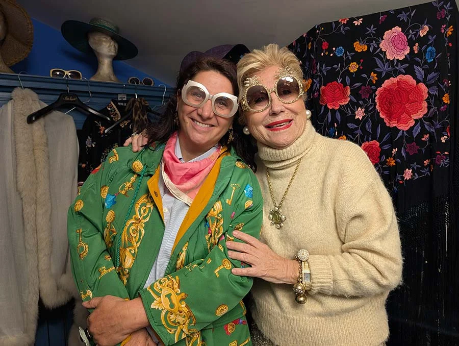 Two smiling women in oversized glasses in a shop: one in a green embroidered jacket, one in a cream jumper and gold pendant