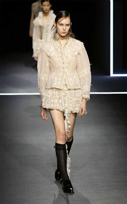 Runway look of a cream lace jacket and matching lace skirt, worn with black knee socks and black shoes