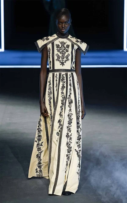 Model in a high-necked cream gown with black embroidered stripes and a floral panel on the bodice, on a dark runway
