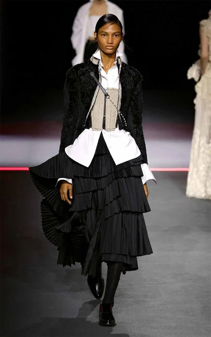 Runway look of a black textured jacket over a white shirt and corset belt, with a tiered pleated black skirt