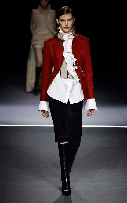 Runway look of a red tailored jacket over a white shirt with a ruffled jabot, black skirt and tall black boots