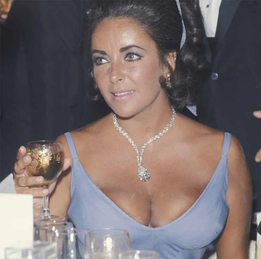 Elizabeth Taylor holding a glass of wine in a pale blue low-cut dress and a diamond pendant necklace
