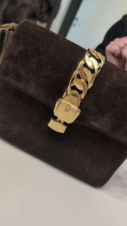 Dark-brown suede vintage Gucci shoulder bag, its gold-tone chain and buckle closure on a table, a hand just behind