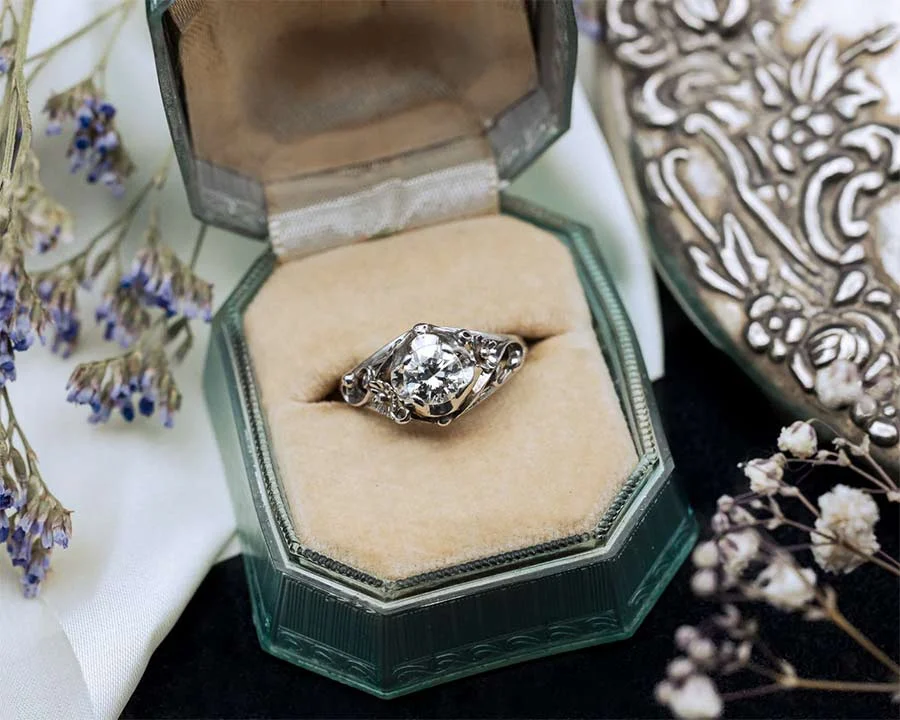 Old-cut diamond solitaire ring with scrolled shoulders, resting in a green-edged vintage ring box beside dried flowers