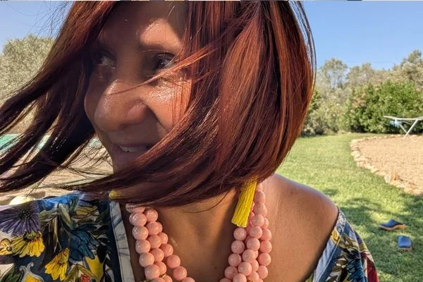 Woman with windblown auburn hair wearing a pink bead necklace and a long yellow tassel earring over a floral top