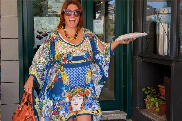 A woman in a blue and yellow majolica-print kaftan, with a Sicilian head-vase motif at the hem, laughing in a shop doorway