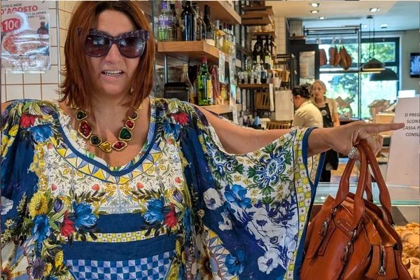 Woman in sunglasses and a floral kaftan with a red and green stone necklace, holding an orange-brown leather bag in a café