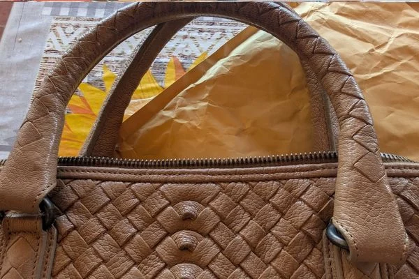 Braided-effect leather handle above the zip and woven body of the bag, against crumpled paper and a patterned cloth