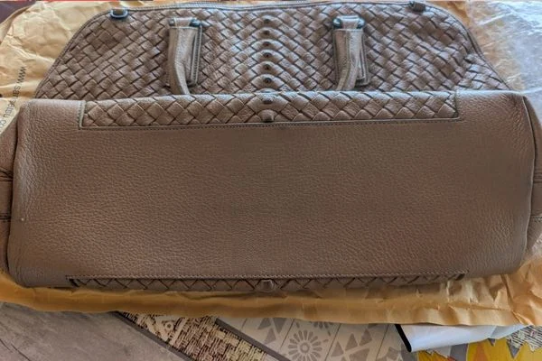Base of the taupe Bottega Veneta bag laid flat on wrapping paper, a smooth grained leather panel below the woven top