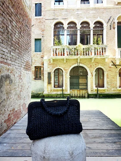 The black woven clutch propped on a stone post, with arched palazzo windows above a green canal