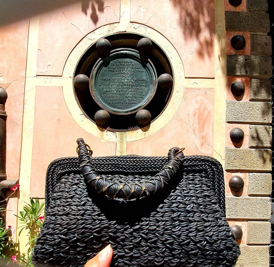 The black woven clutch with its braided handle against the pink wall and round bronze plaque
