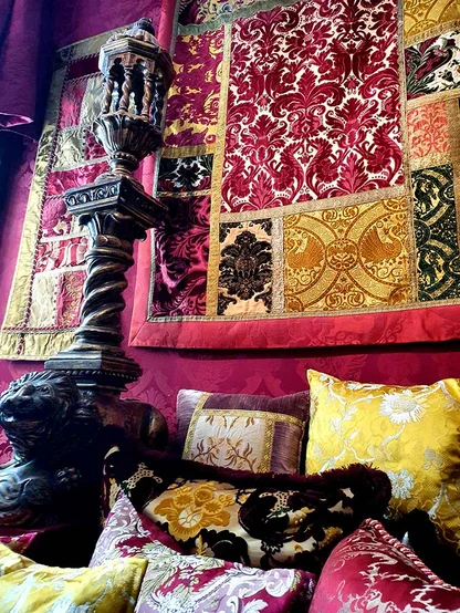 A wall hung with crimson and gold velvet panels above silk cushions, beside a carved wooden lamp stand