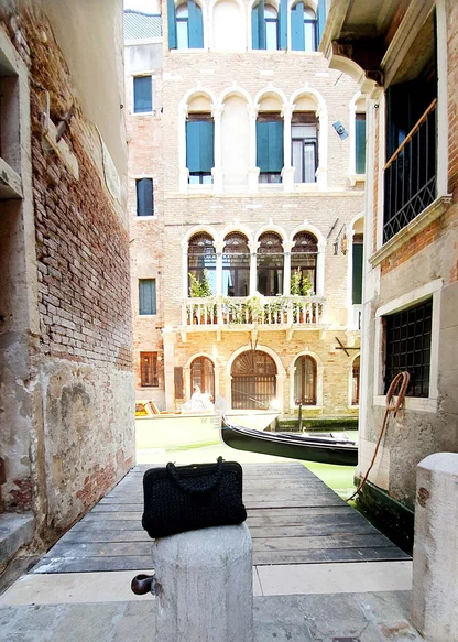 The black woven clutch on a canal-side jetty, with a brick palazzo and a gondola behind