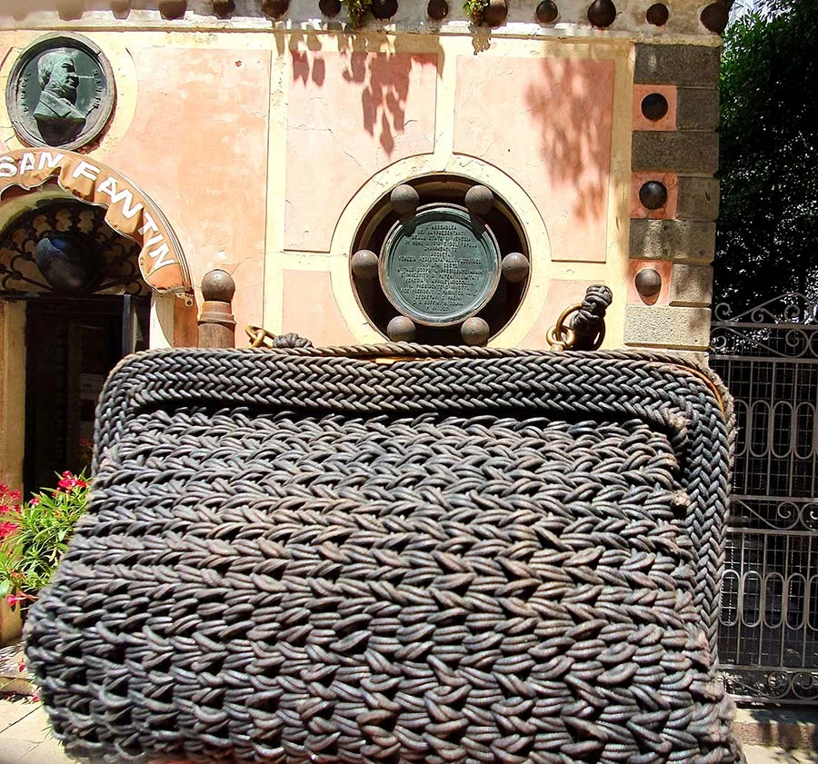 The black woven clutch held up before a pink wall with a round bronze plaque studded with brass balls
