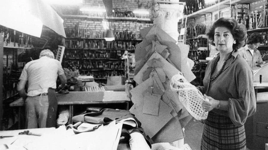 Black-and-white photograph of a woman holding a woven bag in a leather workshop, paper patterns hanging beside her