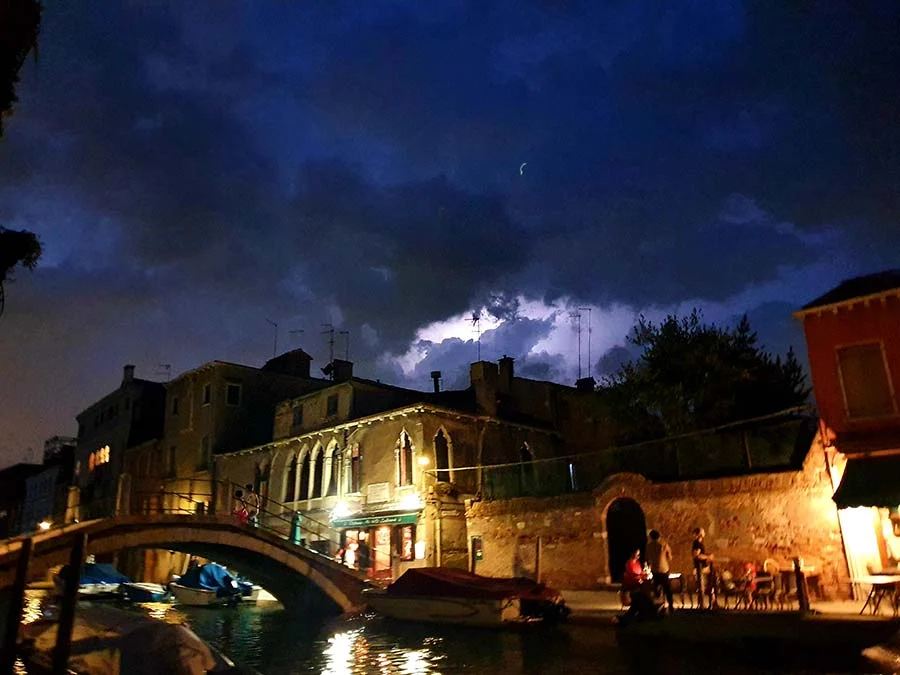Dark storm clouds over a Venice canal at night, with a stone bridge and a lit café terrace