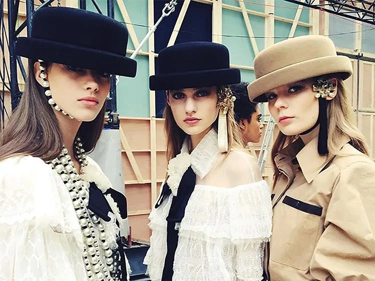 Row of three models in brimmed hats, long pearl strands and a pocketed beige jacket, against a timber backdrop