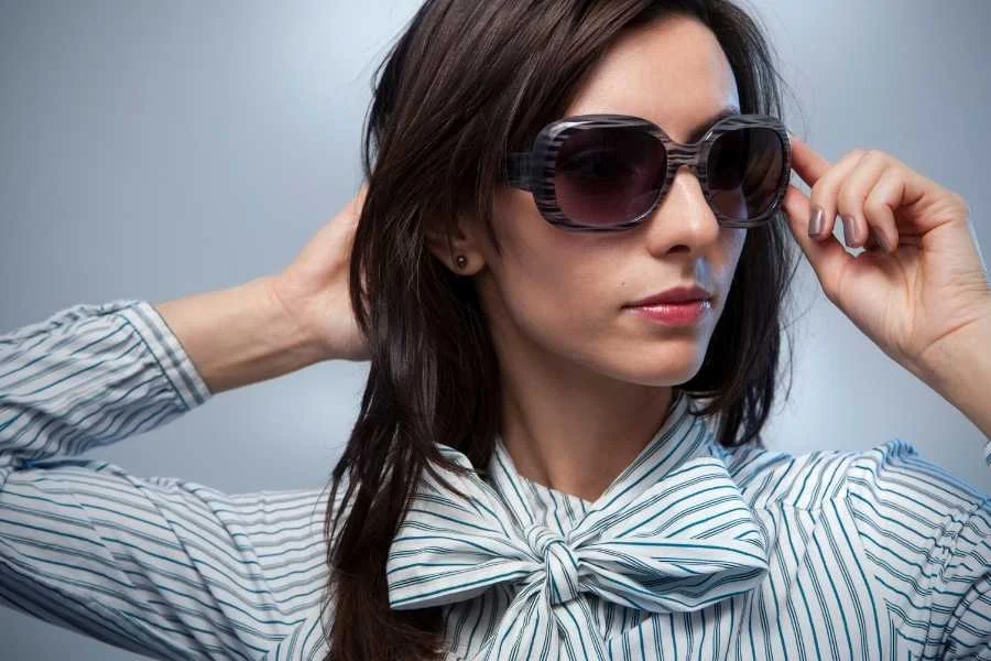 A woman in large grey-striped sunglasses and a blue-striped blouse with a bow at the neck