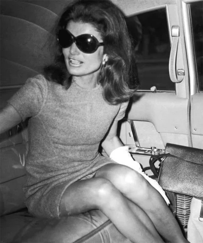 Black-and-white press photograph of Jackie Kennedy climbing out of a car in a short grey dress and oversized sunglasses