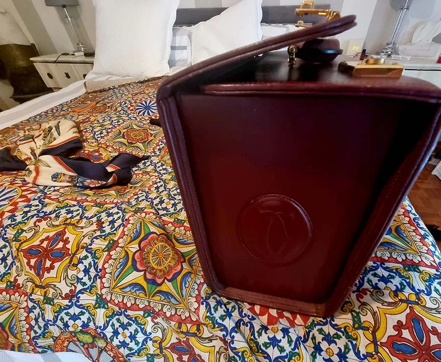 Side of the burgundy Cartier case showing the embossed double C logo, standing on a patterned bedspread